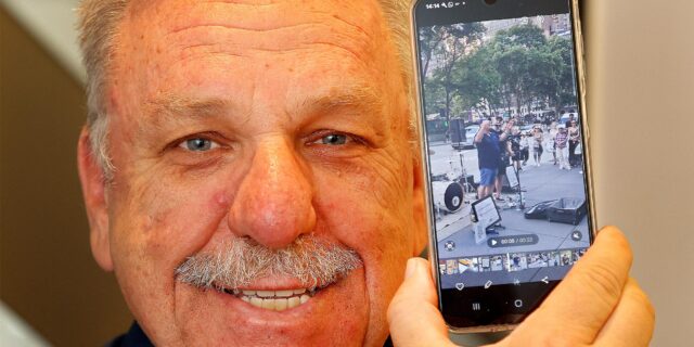 Achim Bauer mit einem Smartphone, das ihn zeigt, als er das Publikum einer Straßenband auf dem Broadway in New York anheizt.