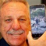 Achim Bauer mit einem Smartphone, das ihn zeigt, als er das Publikum einer Straßenband auf dem Broadway in New York anheizt.