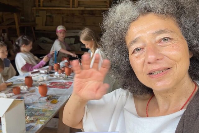 Archäologin und Museumspädagogin Gisela Michel schaut in die Kamera und winkt. Im Hintergrund töpfern Kinder.
