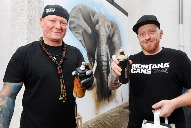 Die Künstler Ted Bartnik und Mike Haze (r.) mit Sprühdosen vor einem Wandbild, das einen Elefanten zeigt.