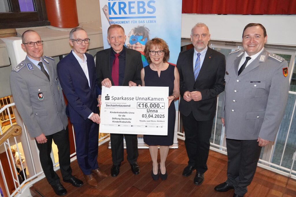 Im Rahmen eines Benefizkonzerts für die Kinderkrebshilfe Unna stehen mehrere Personen in der Stadthalle Unna. Monika und Dieter Rebbert halten einen Scheck