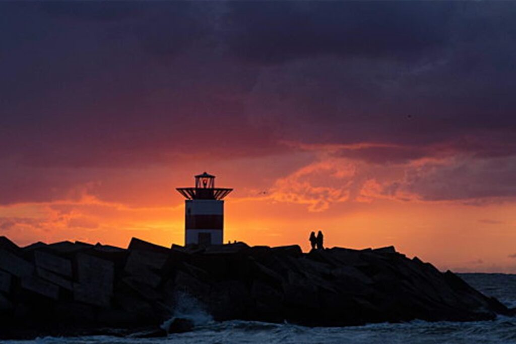 Menschen zeichnen sich als Silhouetten vor dem Abendhimmel ab, während sie in der Nähe eines Leuchtturms am Strand von Scheveningen spazieren. Menschen zeichnen sich als Silhouetten vor dem Abendhimmel ab, während sie in der Nähe eines Leuchtturms am Strand von Scheveningen spazieren.