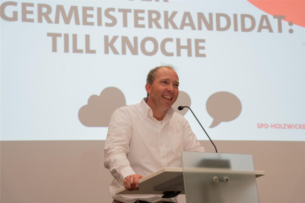 Ein Mann namens Till Knoche Anfang 50 in einem weißen Hemd steht an einem Rednerpult und lächelt. Im Hintergrund ist der Schriftzug Bürgermeisterkandidat: Till Knoche zu lesen. 