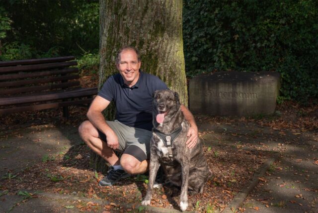 Ein Mann mit dunkelblauem Tshirt hockt neben seinem braun-weißen Hund und lächelt in die Kamera. Im Hintergrund ist eine Parkbank, Laub sowie ein großer Baumstamm zu sehen.