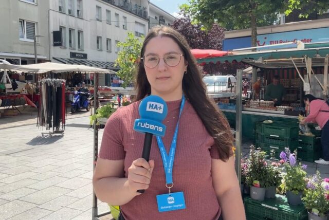 HA-Reporterin Laura Sobczyk mit Mikrofon und blauem Lanyard steht auf dem Wochenmarkt in Kamen. Dort hat sie Menschen auf der Straße zu ihren Urlaubsplänen und Reisen befragt.