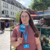 HA-Reporterin Laura Sobczyk mit Mikrofon und blauem Lanyard steht auf dem Wochenmarkt in Kamen. Dort hat sie Menschen auf der Straße zu ihren Urlaubsplänen und Reisen befragt.