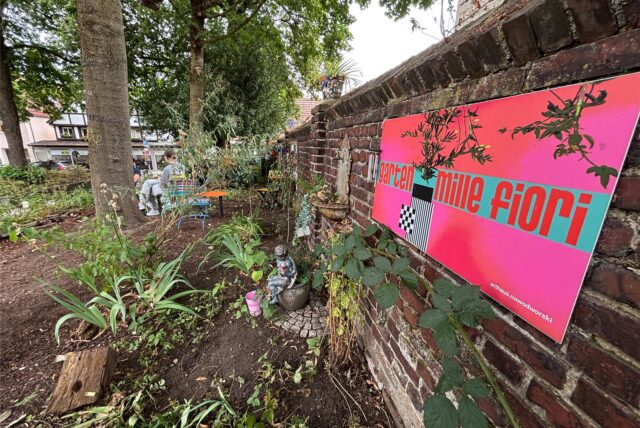Im Garten Mille Fiori in der Innenstadt von Unna wachsen ein paar Pflanzen, an der Mauer hängt ein großes pinkes Schild mit der Aufschrift „Garten Mille Fiori“.
