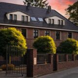 Die Villa wurde aus gebrannten Ziegelsteinen gebaut. Vor der Klinkerfassade sind drei kleiner Bäume und ein Eingangsbereich mit einem schwarzen Tor zu sehen.