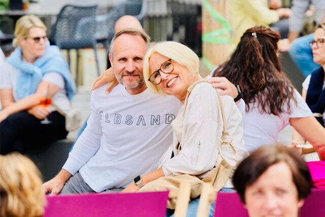 Ein Mann und eine Frau sitzen Arm in Arm beim Feierabendmarkt auf dem Marktplatz in Schwerte. Sie sind von weiteren Menschen umgeben.