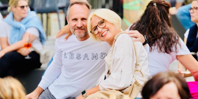 Ein Mann und eine Frau sitzen Arm in Arm beim Feierabendmarkt auf dem Marktplatz in Schwerte. Sie sind von weiteren Menschen umgeben.