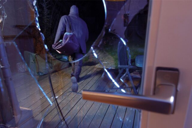Eine schwarz gekleidete Person mit schwarzer Sporttasche läuft im Dunkeln über die Terrasse eines Hauses, im Vordergrund ist eine zerschlagene Fensterscheibe zu erkennen.