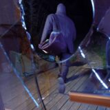 Eine schwarz gekleidete Person mit schwarzer Sporttasche läuft im Dunkeln über die Terrasse eines Hauses, im Vordergrund ist eine zerschlagene Fensterscheibe zu erkennen.
