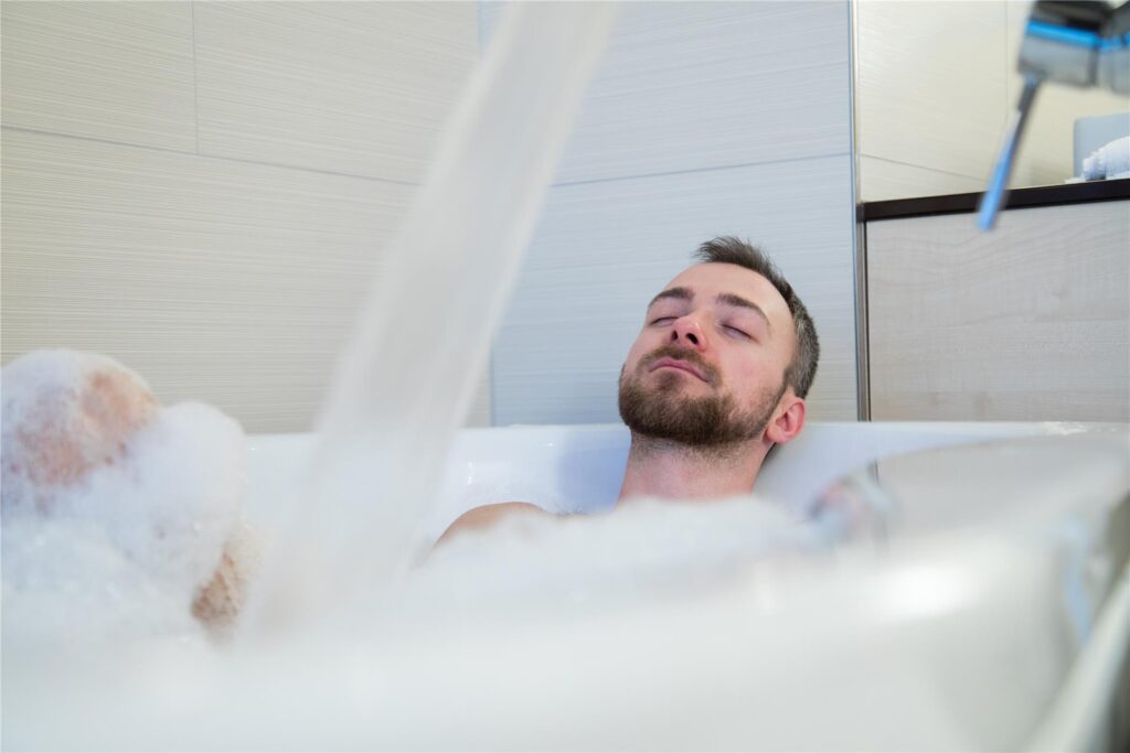 Ein Mann liegt mit geschlossenen Augen in einer gut gefüllten Badewanne. 