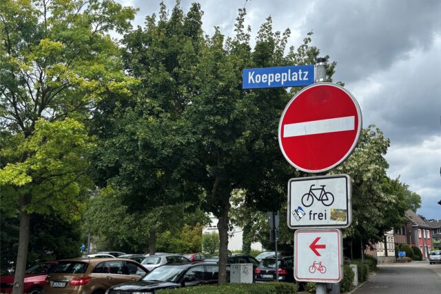 Verkehrsschilder am Koepeplatz in Kamen: Einfahrt verboten, Fahrrad frei, Richtungsweisung für Radfahrer, geparkte Autos und Bäume im Hintergrund. Hier wird es bald zu Baumschnittarbeiten der Stadt kommen.
