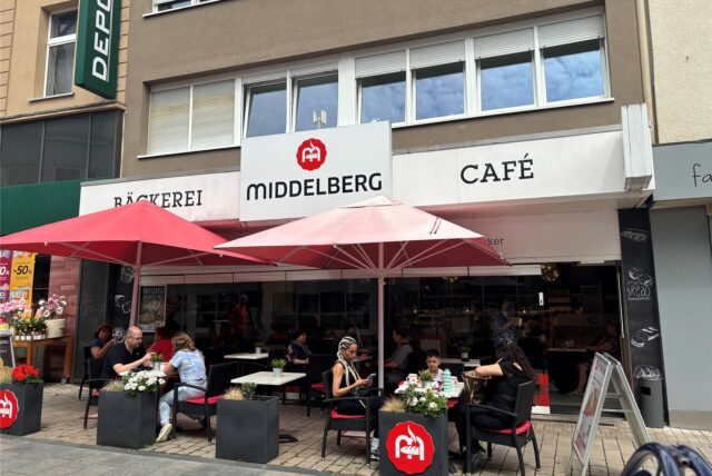 Außenansicht der Bäckerei Middelberg an der Weststraße in Kamen mit Cafébetrieb unter roten Sonnenschirmen – mehrere Gäste sitzen an Tischen vor dem Laden. Am Freitag (18.7.) ist der letzte Verkaufstag.