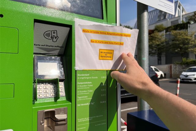 Ein weißes Blatt Papier mit der Aufschrift: „Heute ist leider keine Bargeldzahlung möglich! Nur EC-Karte und Easy Park App. Wir akzeptieren nur Kartenzahlung“. Im Hintergrund sieht man eine Parkfläche mit einzelnen Fahrzeugen.