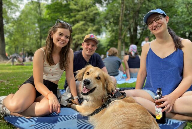 Zwei junge Frauen und ein Mann sitzen auf einer blauen Picknick-Decke im Dortmunder Westpark. Eine der Frauen trinkt ein alkoholfreies Radler. Ebenfalls auf der Decke sitzt ein Hund.