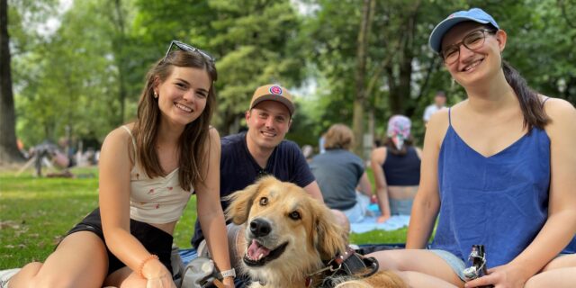 Zwei junge Frauen und ein Mann sitzen auf einer blauen Picknick-Decke im Dortmunder Westpark. Eine der Frauen trinkt ein alkoholfreies Radler. Ebenfalls auf der Decke sitzt ein Hund.