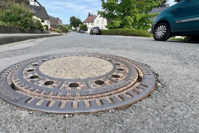Ein Kanaldeckel in der Buschstraße in Unna-Billmerich: Am Straßenrand sind Autos geparkt.