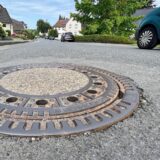 Ein Kanaldeckel in der Buschstraße in Unna-Billmerich: Am Straßenrand sind Autos geparkt.
