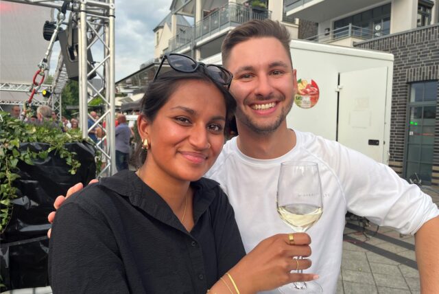 Eine Frau in schwarzer Bluse hält ein Weinglas hoch, ein Mann in weißem Shirt umarmt sie. Beide lächeln in die Kamera.