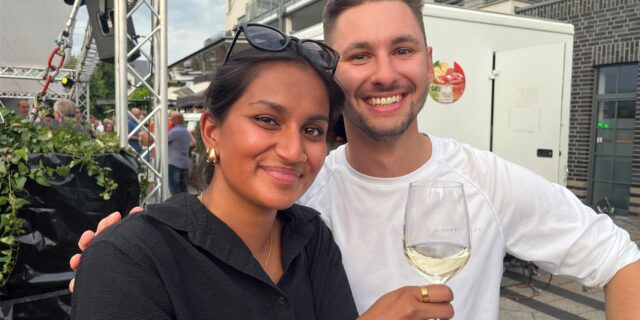 Eine Frau in schwarzer Bluse hält ein Weinglas hoch, ein Mann in weißem Shirt umarmt sie. Beide lächeln in die Kamera.