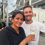 Eine Frau in schwarzer Bluse hält ein Weinglas hoch, ein Mann in weißem Shirt umarmt sie. Beide lächeln in die Kamera.