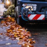 Ein Reinigungsfahrzeug fährt eine Straße entlang und reinigt mit einem Kehrbesen die Straße. Dabei sammelt der Besen Laub auf, dass in einer Linie zusammengefegt wurde.