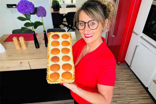 Die Backbloggerin Angie zeigt stolz ihre Aprikosen-Limetten-Tarte. Sie trägt ein rotes Oberteil und eine Brille. Angie lächelt in die Kamera.
