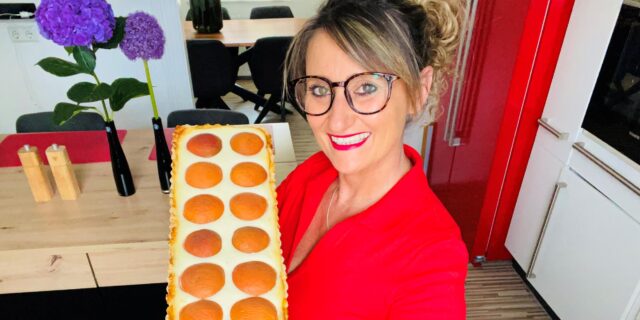 Die Backbloggerin Angie zeigt stolz ihre Aprikosen-Limetten-Tarte. Sie trägt ein rotes Oberteil und eine Brille. Angie lächelt in die Kamera.