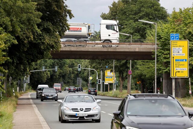 Straßenszene in Kamen mit mehreren Autos auf der Heerener Straße unter der B233-Brücke, auf der ein LKW mit Anhänger fährt; Verkehrsschilder und Ampeln sind sichtbar, umgeben von Bäumen.