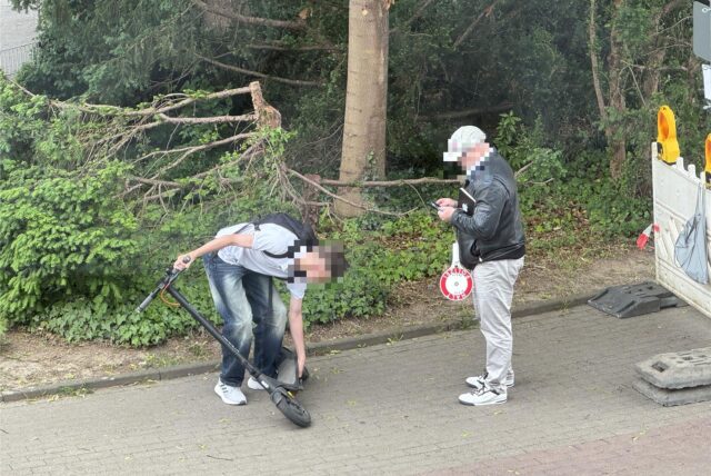 Ein Jugendlicher hantiert an einem E-Scooter, während ein Zivilpolizist daneben steht und etwas notiert. Der Vorfall ereignete sich auf einem Gehweg am Ostring in Unna, im Hintergrund ist Bewuchs zu erkennen.