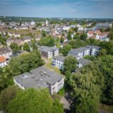 Eine Luftaufnahme zeigt eine Wohngegend mit Mehrfamilienhäusern und Einfamilienhäusern in Dortmund-Hombruch. Die Gebäude sind von viel Grün umgeben, und im Hintergrund ist eine Kirchturmspitze zu sehen.