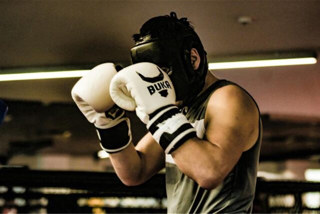 Ein Boxer mit schwarzem Kopfschutz hält seine weißen Boxhandschuhe vor dem Gesicht.