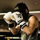Ein Boxer mit schwarzem Kopfschutz hält seine weißen Boxhandschuhe vor dem Gesicht.