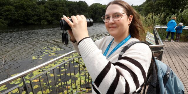HA-Reporterin Laura Sobczyk hält ein Fernglas in der Hand auf der Aussichtsplattform am Beversee in Bergkamen. Sie trägt einen Erlebnisrucksack des Stadtmarketings Bergkamen auf dem Rücken.