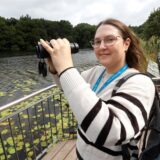 HA-Reporterin Laura Sobczyk hält ein Fernglas in der Hand auf der Aussichtsplattform am Beversee in Bergkamen. Sie trägt einen Erlebnisrucksack des Stadtmarketings Bergkamen auf dem Rücken.