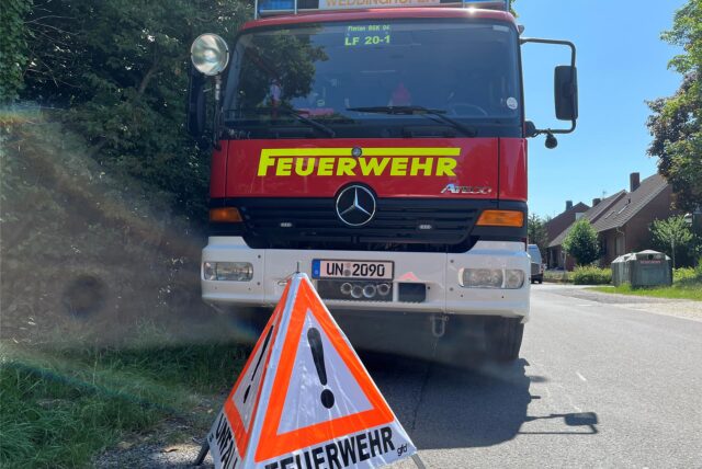 Ein Feuerwehrfahrzeug der Einheit Weddinghofen der Freiwilligen Feuerwehr Bergkamen parkt an der Nördlichen Lippestraße in Heil. Davor steht ein Warndreieck mit der Aufschrift Feuerwehr und einem Ausrufezeichen.