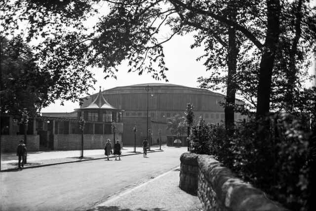 Passanten gehen auf einer Straße in Dortmund in Richtung der alten Westfalenhalle, die im Hintergrund zu sehen ist.