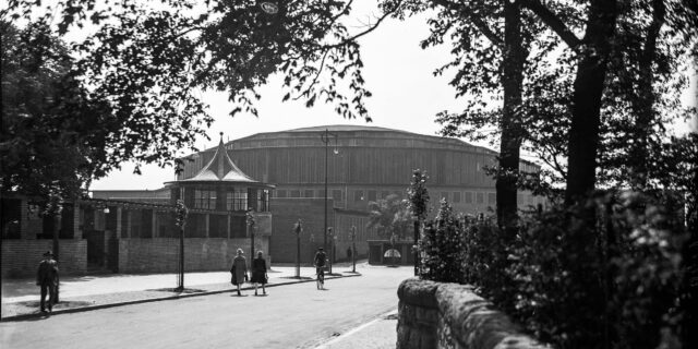 Passanten gehen auf einer Straße in Dortmund in Richtung der alten Westfalenhalle, die im Hintergrund zu sehen ist.