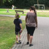 Eine Frau und ein Junge gehen auf einem gepflasterten Weg im Kurpark in Unna-Königsborn. Der Junge trägt gelbe Kopfhörer. Im Hintergrund sind Spielgeräte und andere Parkbesucher zu sehen.