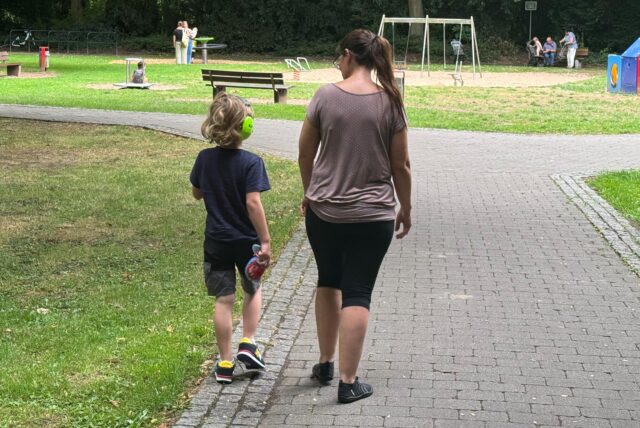 Eine Frau und ein Junge gehen auf einem gepflasterten Weg im Kurpark in Unna-Königsborn. Der Junge trägt gelbe Kopfhörer. Im Hintergrund sind Spielgeräte und andere Parkbesucher zu sehen.