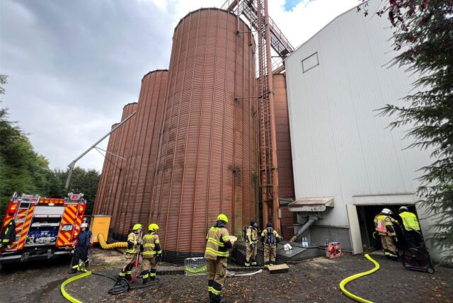 Mehrere Einsatzkräfte der Feuerwehr vor den Silos der Mühle Sendker in Kamen. Der neongelbe Feuerwehrschlauch ist ausgerollt.