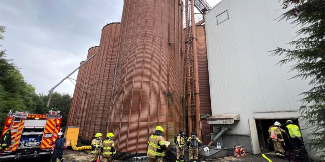 Mehrere Einsatzkräfte der Feuerwehr vor den Silos der Mühle Sendker in Kamen. Der neongelbe Feuerwehrschlauch ist ausgerollt.