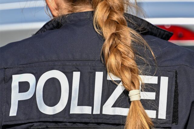Das Bild zeigt eine Polizistin von hinten. Sie hat einen geflochtenen Zopf und auf ihrer Jacke steht in großen weißen Buchstaben POLIZEI.