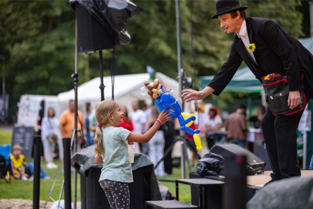 Comedia Tobi van Deisner übergibt einem kleinen Mädchen aus dem Publikum eine Luftballon-Puppe beim Kulturpicknick im Römerpark in Bergkamen.
