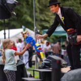 Comedia Tobi van Deisner übergibt einem kleinen Mädchen aus dem Publikum eine Luftballon-Puppe beim Kulturpicknick im Römerpark in Bergkamen.